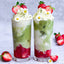 Two tall glasses of matcha latte layered with strawberries and whipped cream on a light background